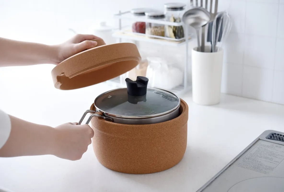 清潔なキッチンで、コルク製の保温容器に入った鍋の蓋を開け閉めしている場面。奥には調味料や調理器具が整然と並び、料理の準備中の日常風景がうかがえます。