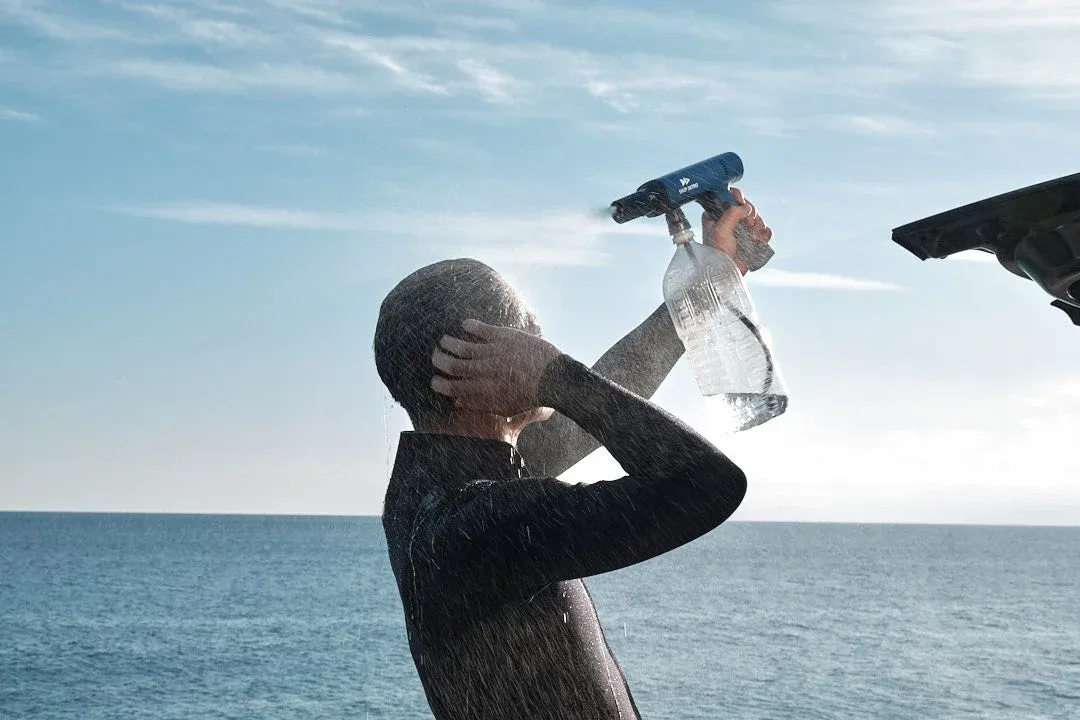 ウェットスーツを着た人が、海を背景にポータブルシャワーを使って体を洗い流している様子