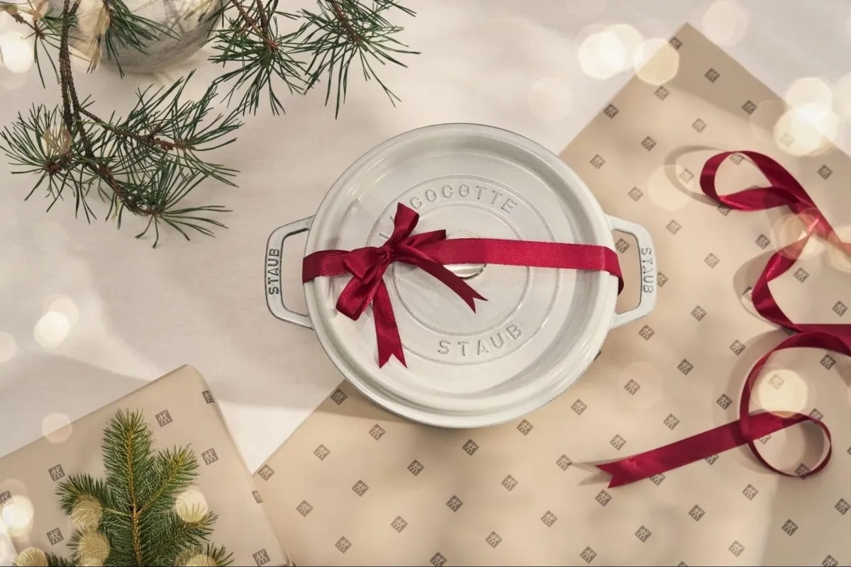 白いストウブのココット鍋が赤いリボンで飾られ、クリスマスらしいラッピングペーパーと松の枝と共に写っています。ホリデーシーズンの贈り物にぴったりの、温かみのある雰囲気です。