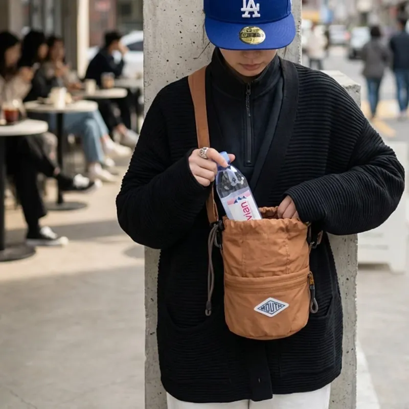 青いキャップと黒いカーディガンを着た人物が、エビアンのペットボトルを茶色のショルダーバッグに入れている様子