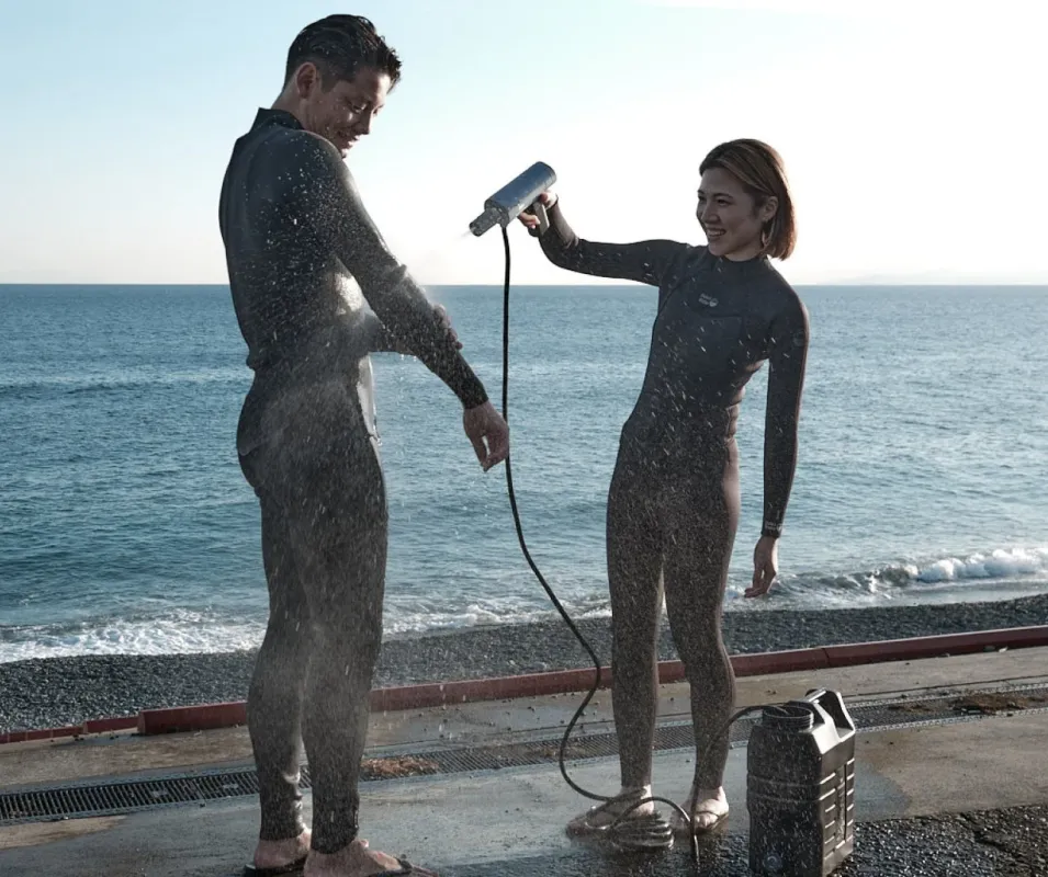 ウェットスーツを着た男女が海辺でポータブルシャワーを使って体を洗い流している様子