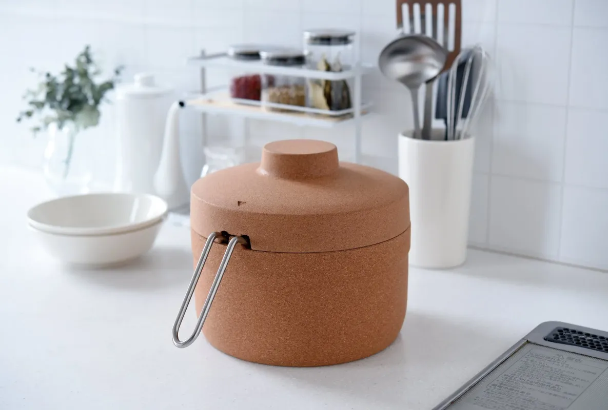 白いタイル張りの清潔感あるキッチンカウンターに、コルク調のユニークなポットが置かれている。背景にはスパイスラックや調理器具が見え、モダンで機能的な台所の雰囲気を醸し出している。