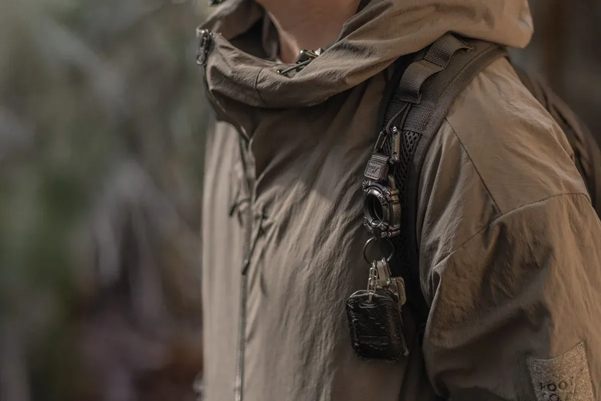 カーキ色のフード付きジャケットを着た人物が、バックパックのショルダーストラップにタクティカルなキーホルダーと鍵を付けている