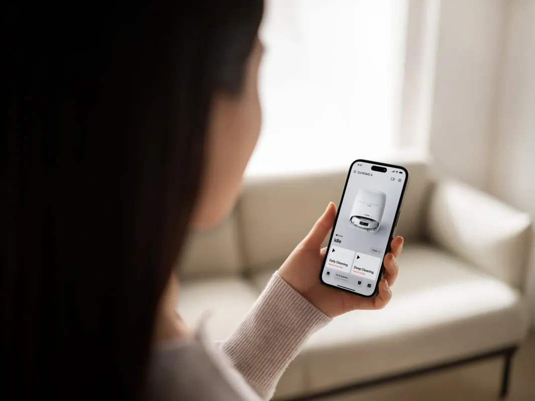 女性がスマートフォンを操作し、ロボット掃除機の管理アプリ画面を見ている。リビングでスマート家電を活用する現代的なライフスタイルを表現している。