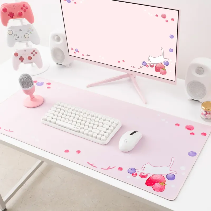 ピンクを基調とした可愛らしいゲーミングデスクのセットアップ。猫といちごのイラストが描かれたモニターとマウスパッドが特徴的で、白いキーボード、マウス、ピンクのコントローラーなどが配置されている。
