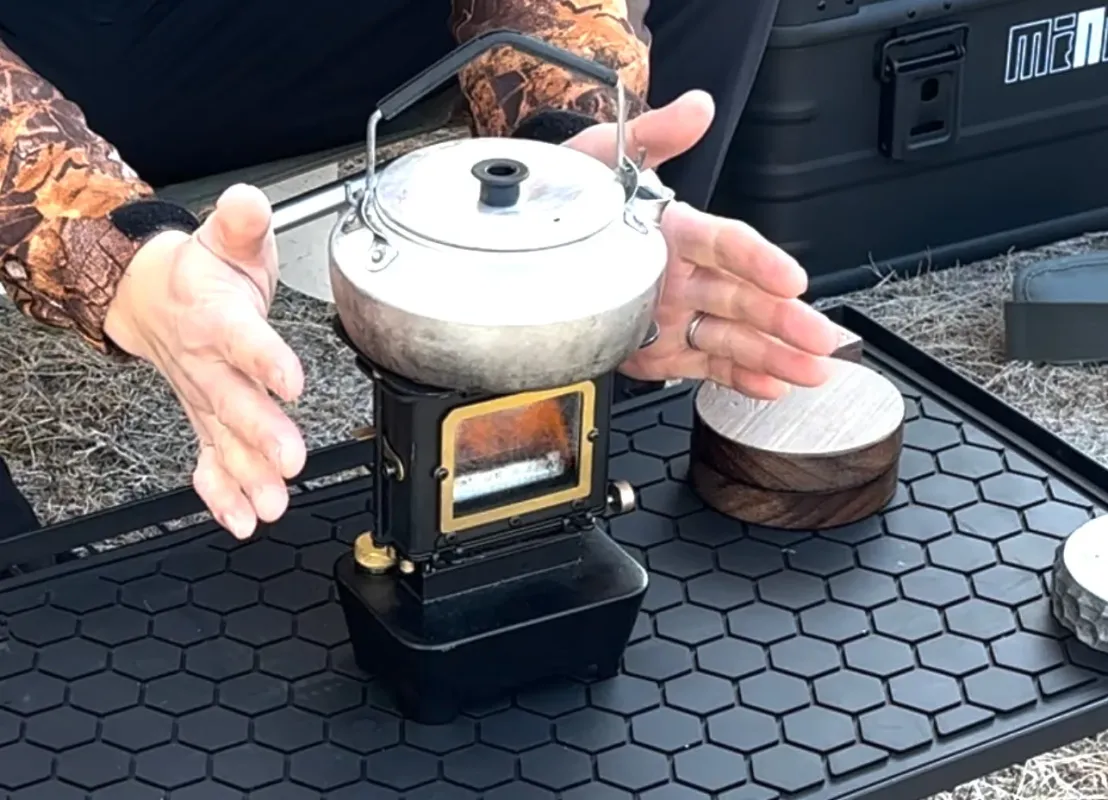 迷彩柄の服を着た人が、小型のポータブルストーブでケトルを温めている様子