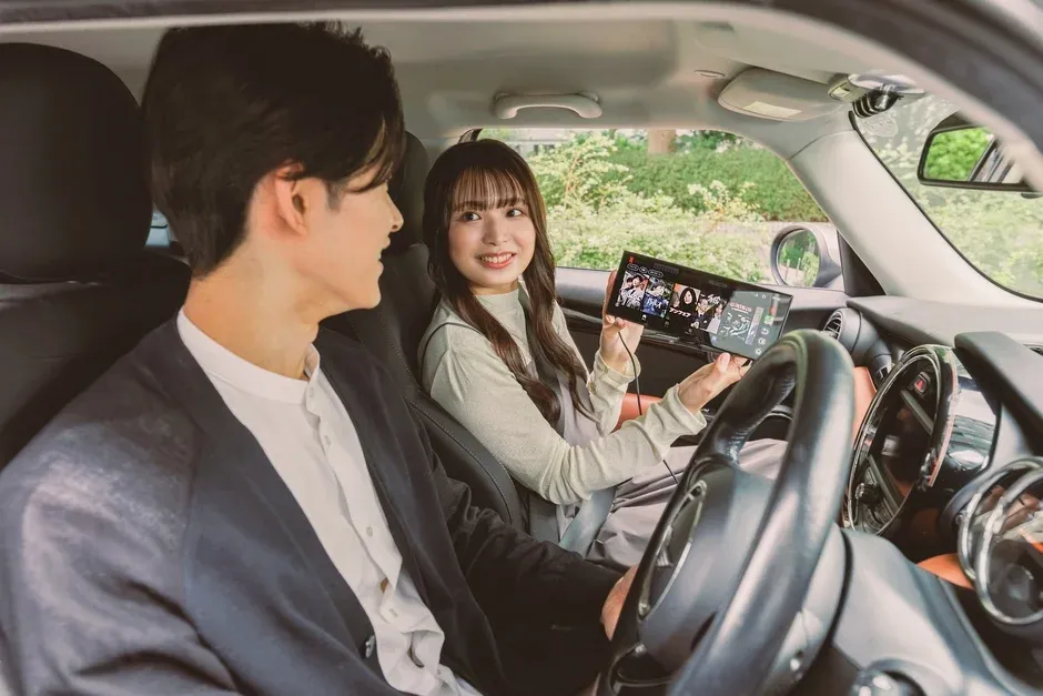 車内でくつろぐ若いカップルが、助手席の女性が持つタブレット端末の画面を共有し、笑顔でコンテンツを視聴している様子
