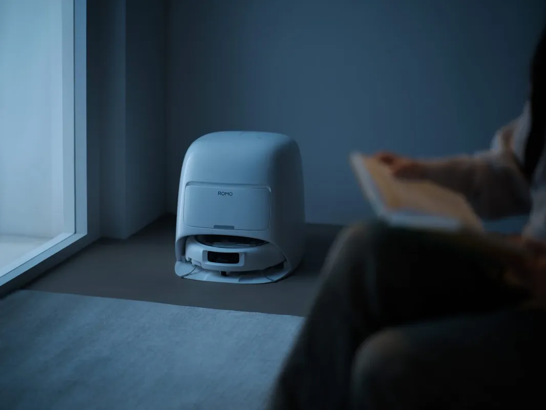 夜の室内で、白いロボット掃除機がドッキングステーションに収まっており、その手前で人物が読書をしている様子。静かで落ち着いた雰囲気を感じさせます。