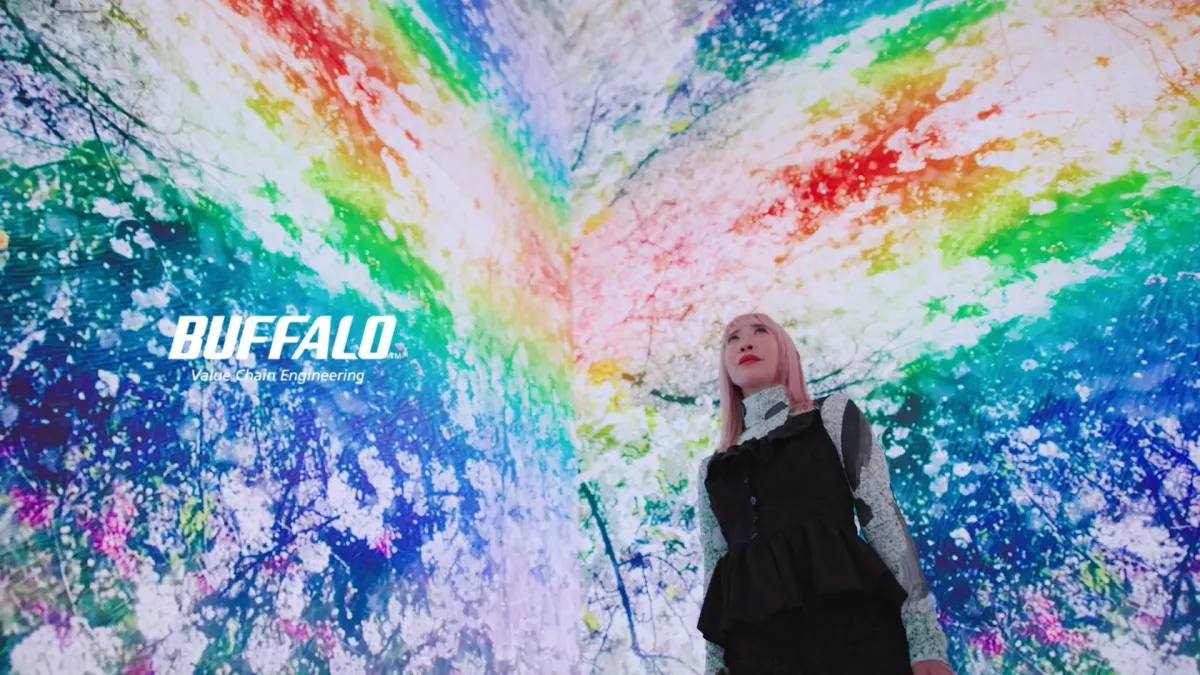 ピンク髪の女性が、桜のような模様と虹色のグラデーションが広がる没入型デジタルアート空間を見上げている様子