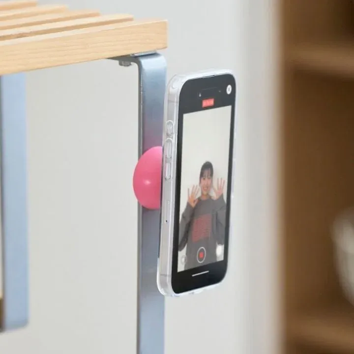 ピンクの磁石で金属製の棚にスマートフォンが固定され、画面に笑顔の女性が映る