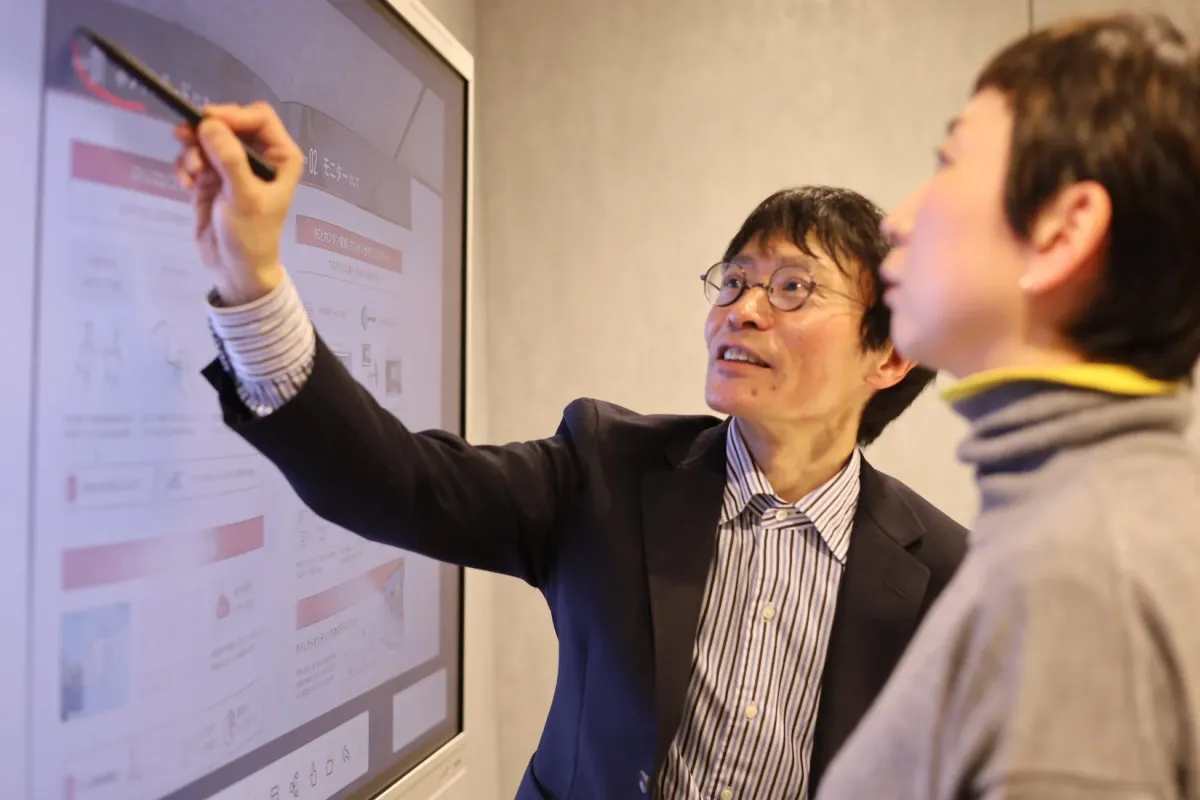 男性が大型モニターを指し示しながら女性に何かを説明しているビジネスシーン