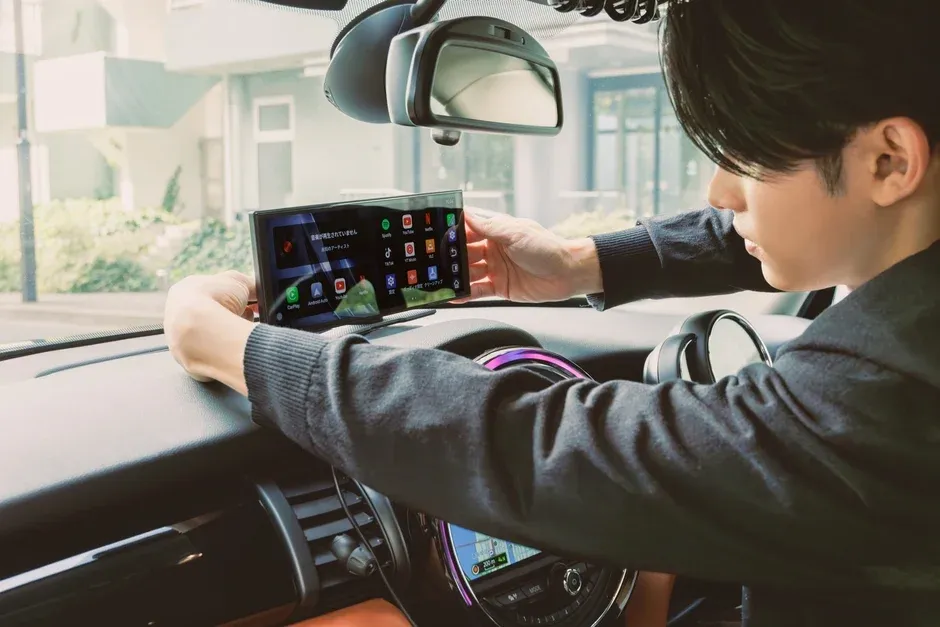 車内で男性がダッシュボード上のポータブルディスプレイを操作しており、SpotifyやYouTubeなどのアプリアイコンが表示されている