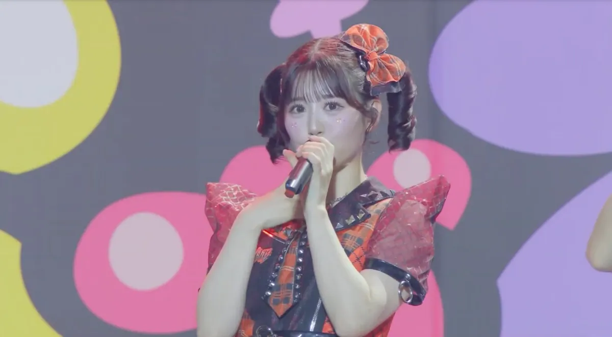 ステージ上でマイクを握り歌唱する、チェック柄の衣装とツインテールが特徴的な女性アイドル