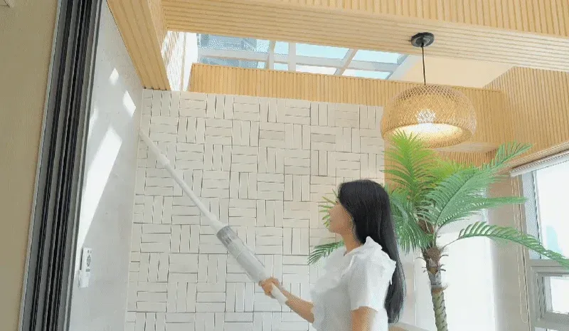 明るくモダンなリビングで、女性が白いスティック型クリーナーを使って壁を掃除している様子。