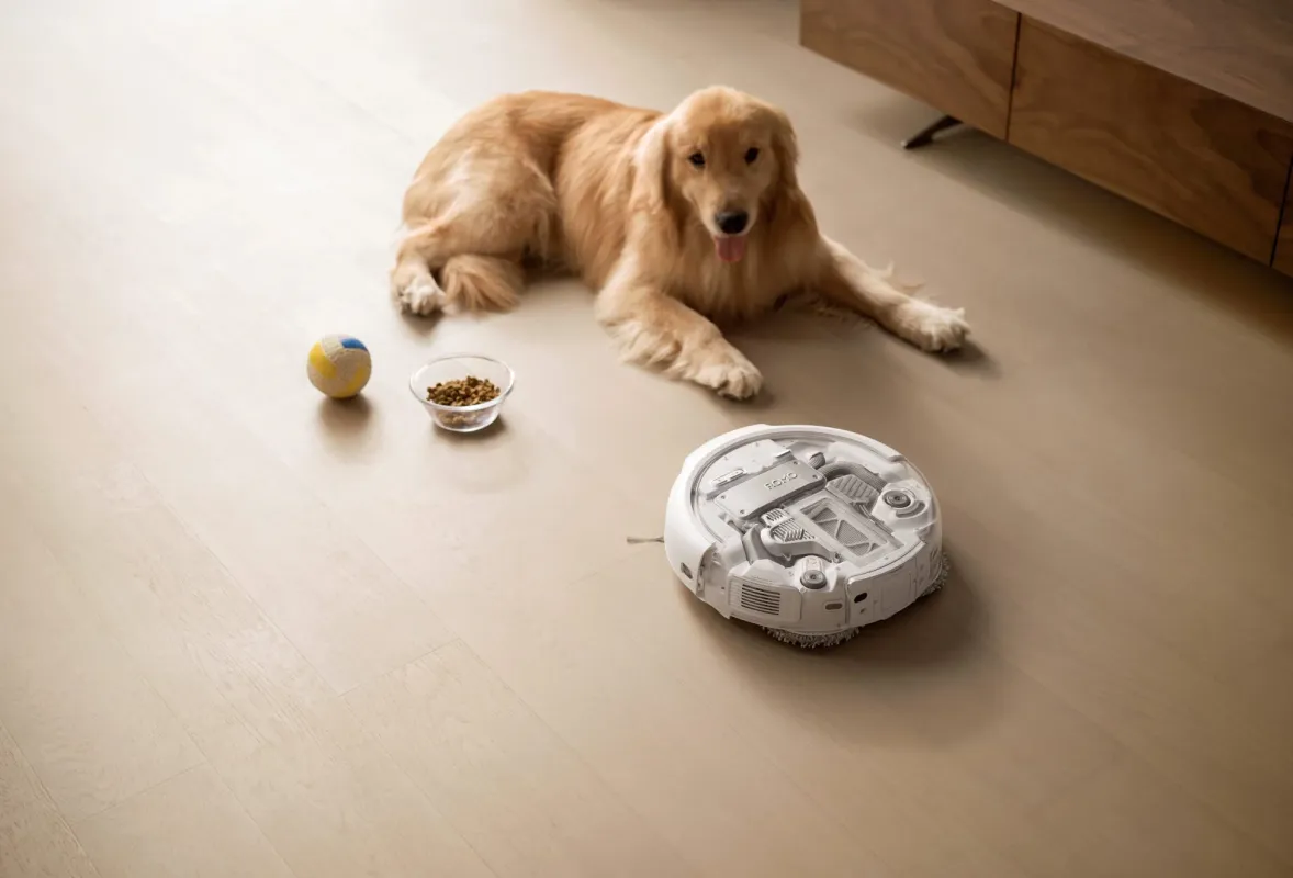 ゴールデンレトリバーがフローリングの部屋でくつろいでいる様子。そばにはドッグフードのボウル、ボール、そして白いロボット掃除機が置かれており、ペットと家電が共存する穏やかな日常風景を捉えています。
