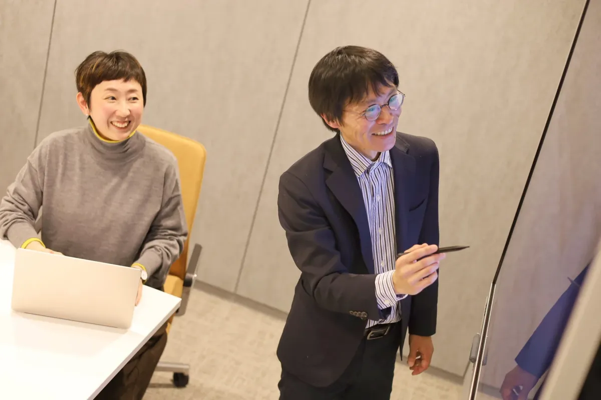 オフィスで、男性がデジタルホワイトボードを指しながらプレゼンテーションをしており、女性が笑顔で話を聞いている様子