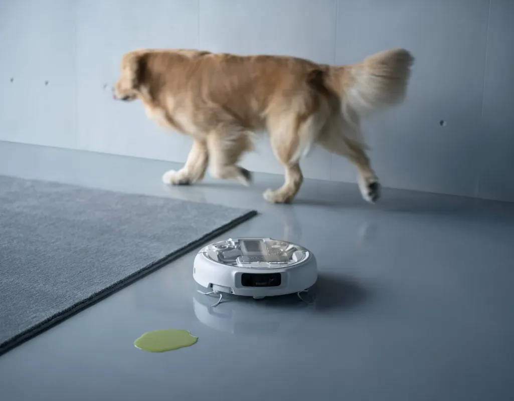 ゴールデンレトリバーが歩く室内で、ロボット掃除機の近くに緑色の液体がこぼれています。ペットとスマート家電のある現代的な生活空間における清掃の課題を示唆しています。