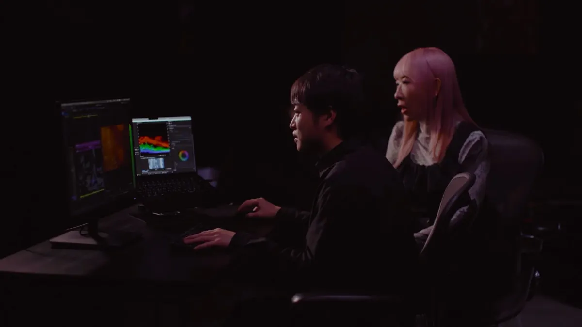 暗い部屋で男女2人がPC画面を見つめ、映像制作やカラーグレーディング作業を行っている様子