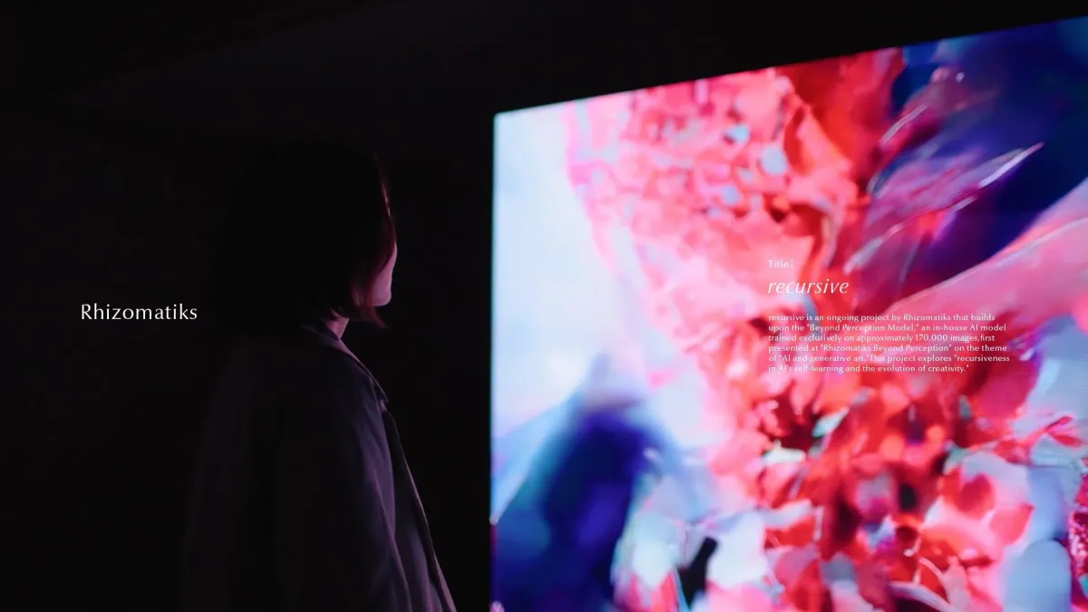 暗い空間で一人の人物が、鮮やかなピンクと青の抽象的なジェネレーティブアートが映し出された大型スクリーンを鑑賞