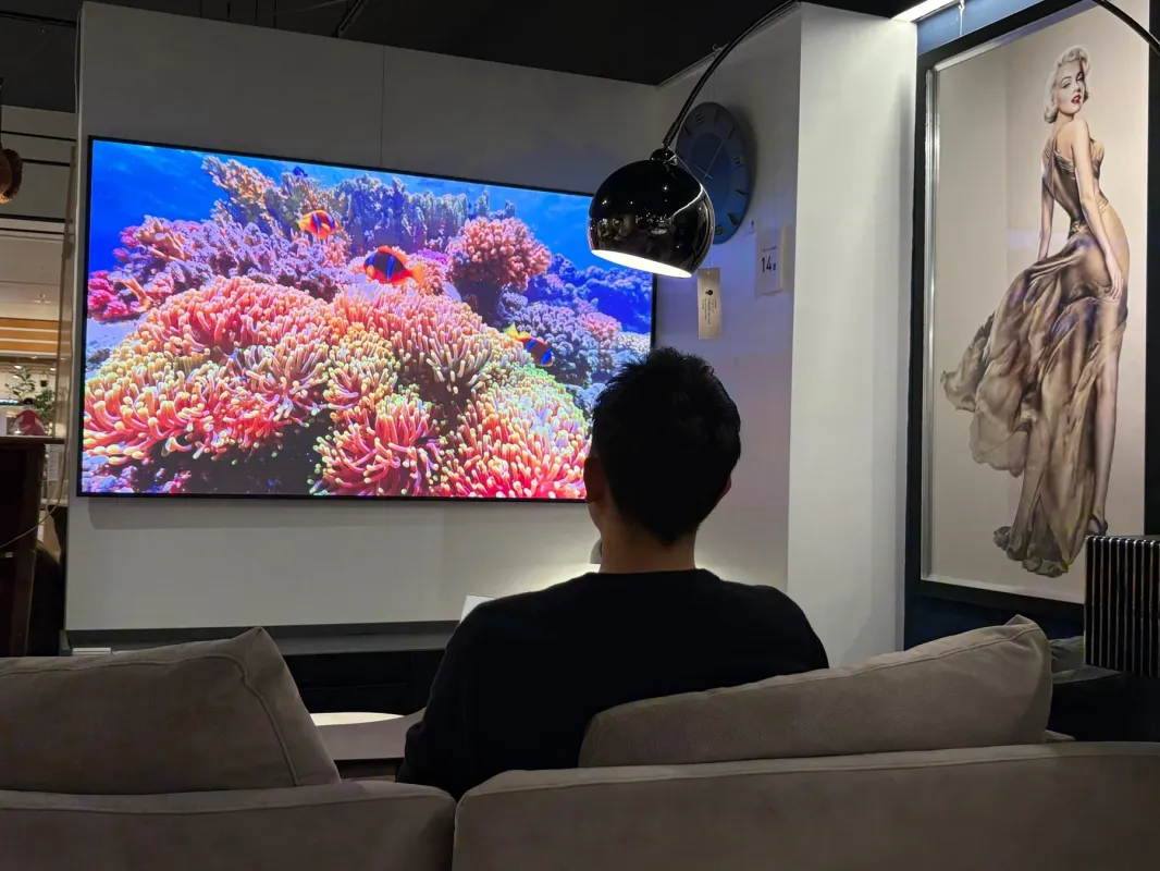 ソファに座った男性が、鮮やかなサンゴ礁とカクレクマノミが映る大型テレビを鑑賞している。モダンなリビングで、リラックスしてエンタメを楽しむ様子が伺える。