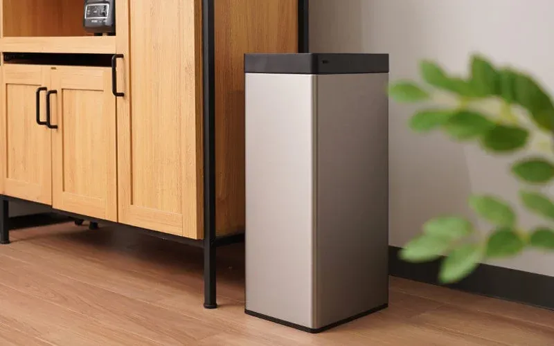 銀色のモダンなゴミ箱が、木製のキャビネットと観葉植物のある室内に置かれている様子。フローリングの床と白い壁が背景にあり、シンプルで整頓された空間を演出しています。