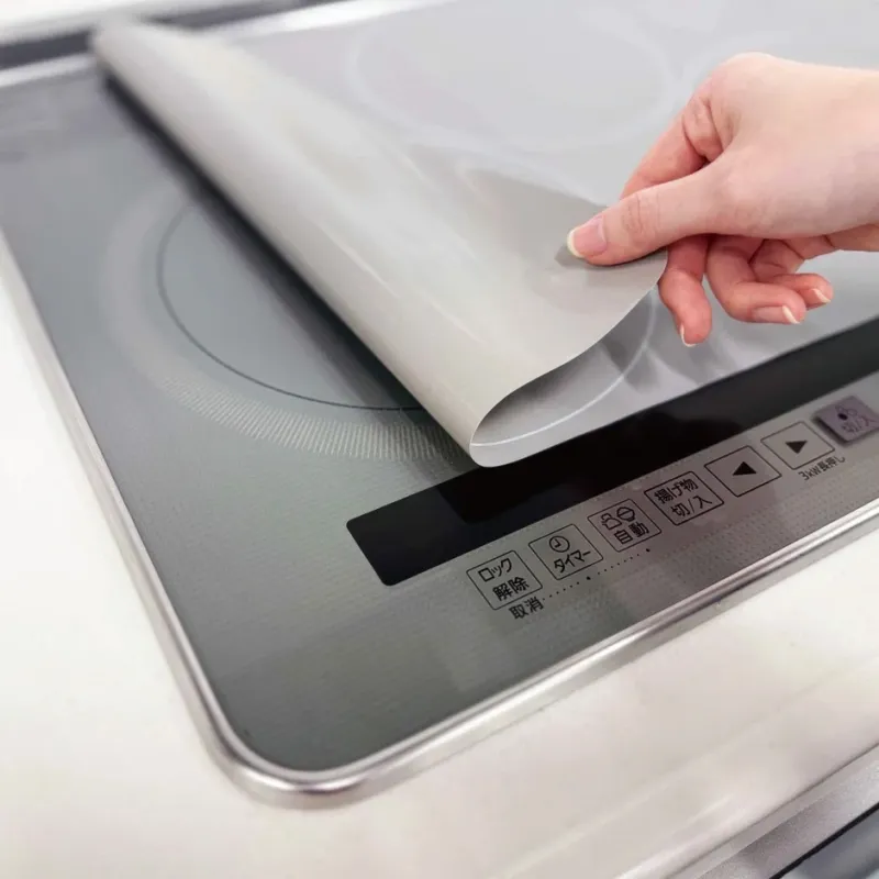 IHクッキングヒーターの天板に貼られた保護シートを剥がしている手の様子です。調理面を傷や汚れから守るためのシートで、清潔感を保つためのアイテムであることが伺えます。