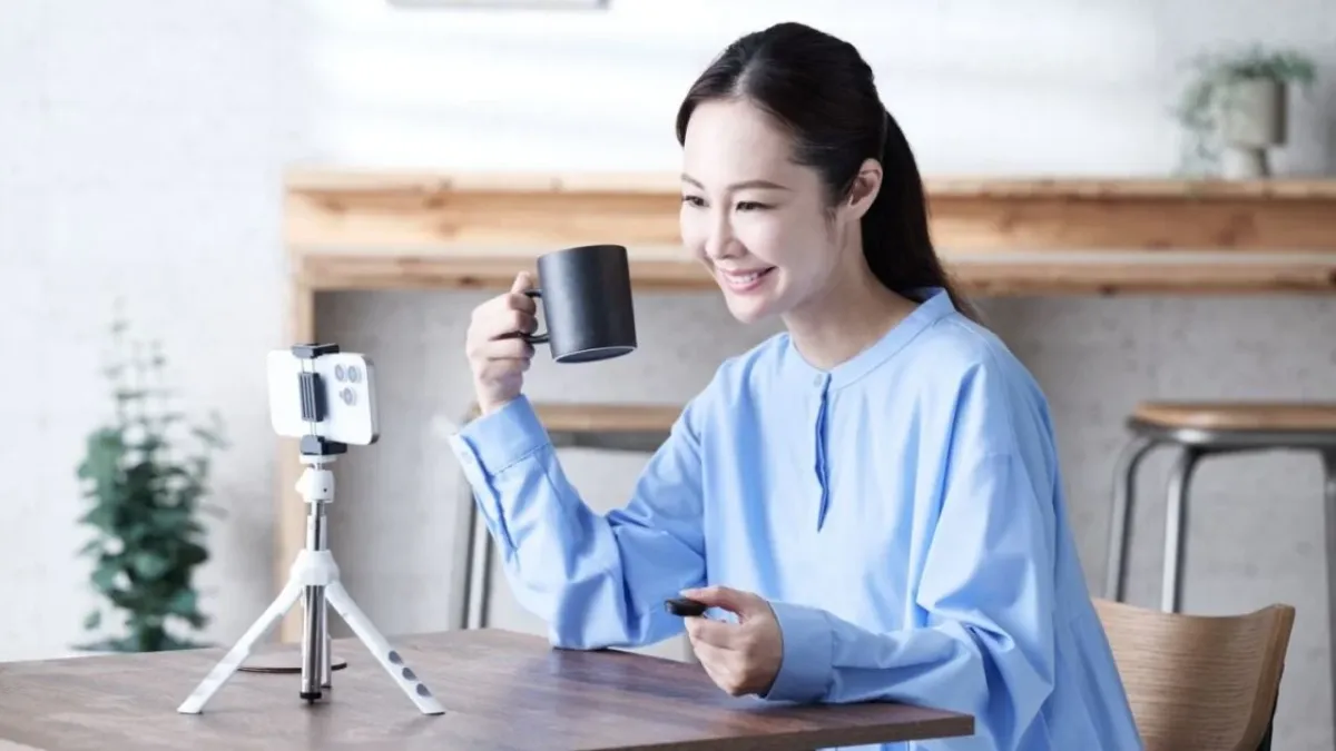 笑顔の女性が自宅のような場所で、スマートフォンを三脚にセットしてライブ配信やビデオ通話をしている様子