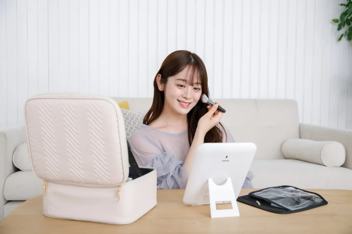 笑顔の若い女性がメイクアップブラシを使って化粧している。メイクポーチとミラーがテーブルに。