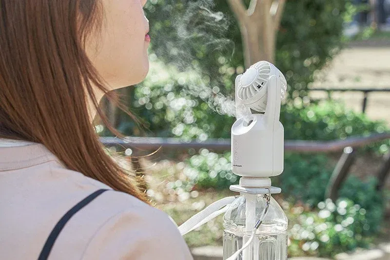 屋外で女性がポータブルミストファンを使用し、顔に涼しいミストを浴びている様子。ペットボトルに装着されたデバイスからミストが噴き出しており、夏の暑さ対策に効果的です。