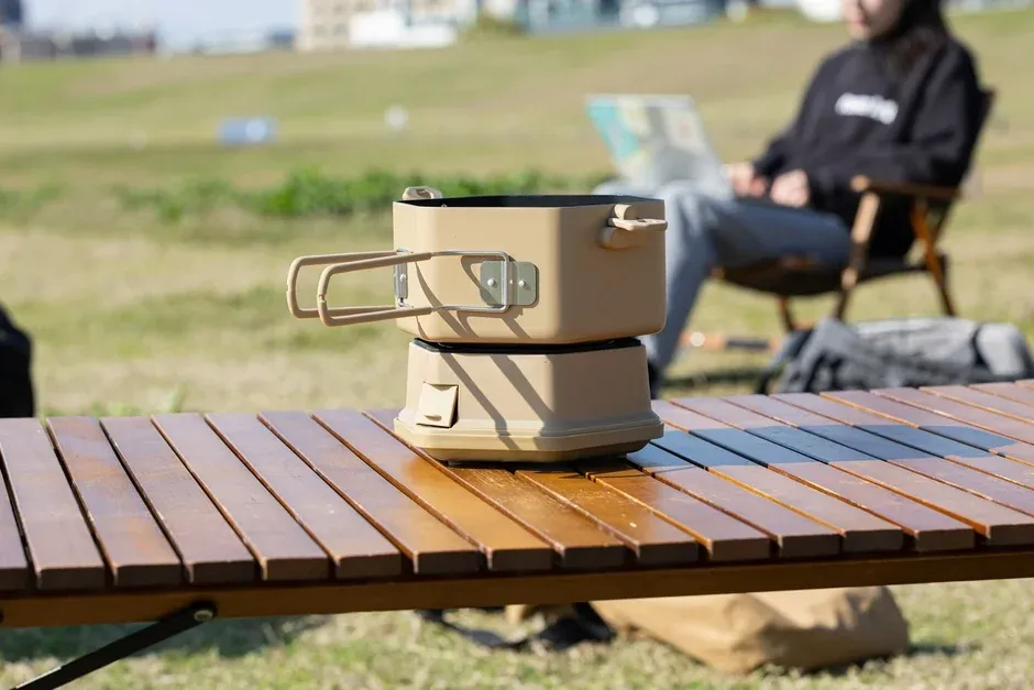 青空の下、キャンプ用テーブルに置かれたクッカーが手前にあり、奥では人物が椅子に座ってくつろいでいるアウトドアシーン
