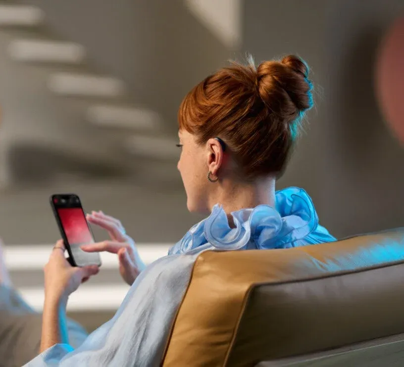 赤い髪をシニヨンにまとめた女性が、後ろ姿でソファに座りスマートフォンを操作している様子