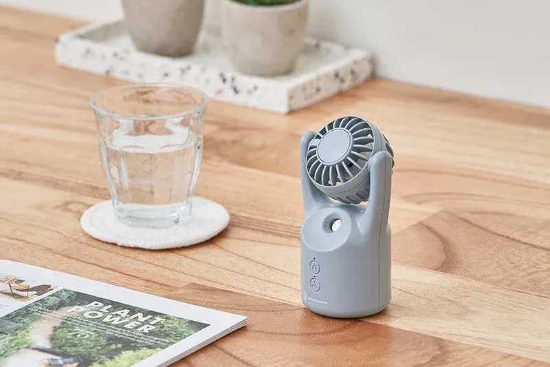 木目のテーブルに置かれたグレーのポータブル扇風機と水の入ったグラス、雑誌が写っています。涼しげで快適な夏のひとときを連想させる、落ち着いた雰囲気の画像です。