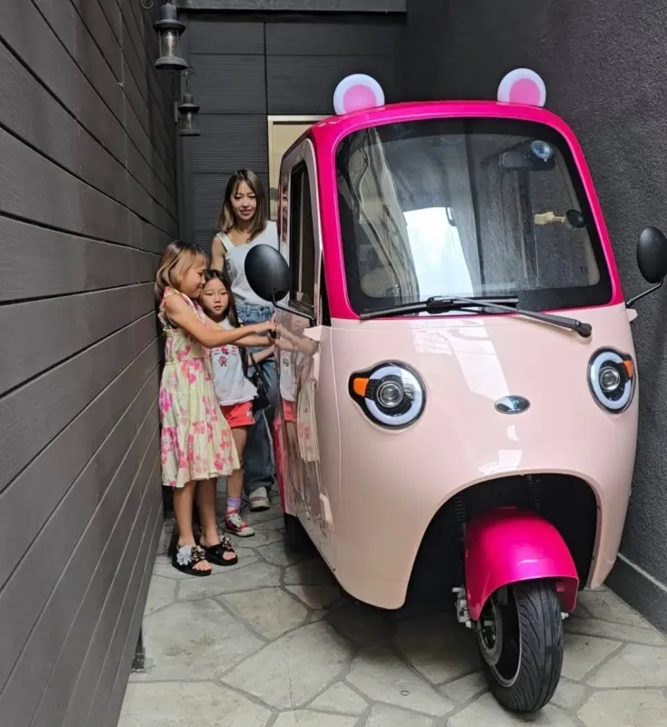 ピンク色のクマ耳付き三輪車と、それを見つめる女性と二人の子供たち