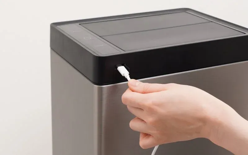 メタリックな筐体の電子機器に、白いUSBケーブルを接続している手のクローズアップ。デバイスの黒い上部には操作パネルのようなものが見えます。